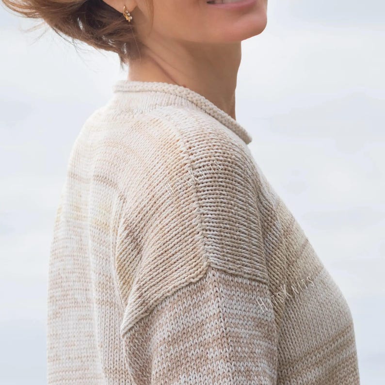 May include: A beige and white marled knit sweater featuring a crew neckline and drop shoulder seams. The garment is crafted with a textured stitch pattern and includes the subtle text "HUSK KNITS" printed on the upper right chest area.