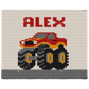 May include: A rectangular knit fabric piece featuring the name ALEX in red block letters above a graphic of a red and orange monster truck with large black tires. The background is cream-colored with a solid dark gray border at the bottom.