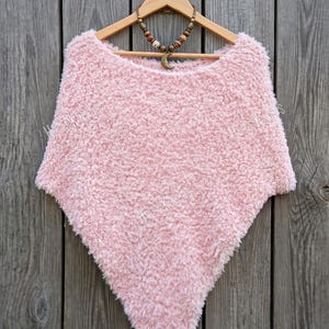May include: A light pink, fuzzy, triangular-shaped poncho made of soft, textured yarn. The garment is displayed on a wooden hanger and features a wide neckline. A beaded necklace with a crescent moon pendant rests on the hanger above the poncho.