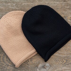 May include: Two knitted beanies, one black and one beige, are lying on a wooden surface.