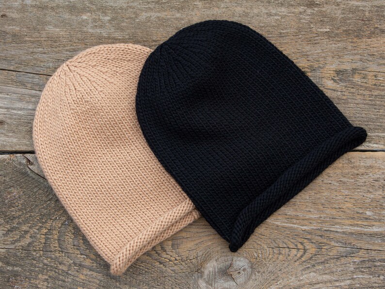 May include: Two knitted beanies, one black and one beige, are lying on a wooden surface.