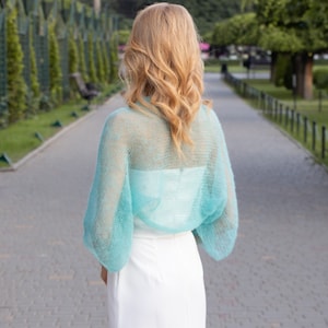 May include: A light blue sheer knitted shrug with long sleeves. The shrug is made of a delicate, airy fabric and has a loose, flowing fit.
