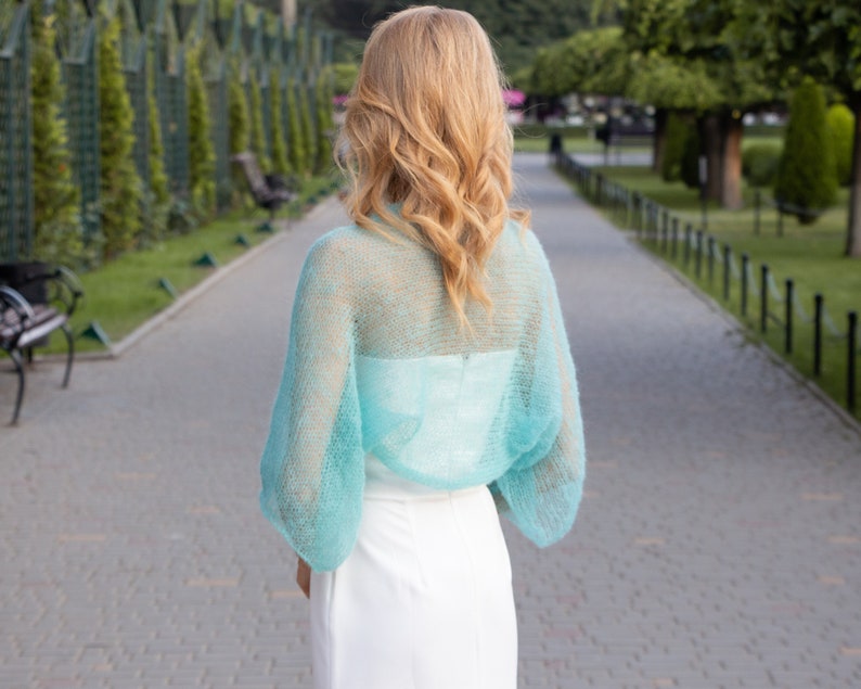 May include: A light blue sheer knitted shrug with long sleeves. The shrug is made of a delicate, airy fabric and has a loose, flowing fit.