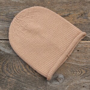 May include: A plain, light brown knitted beanie hat on a rustic wooden background.