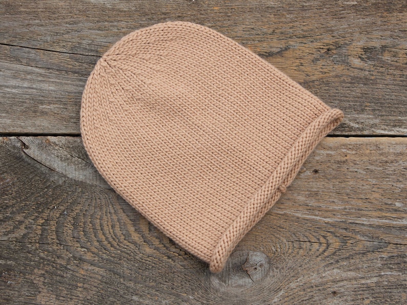 May include: A plain, light brown knitted beanie hat on a rustic wooden background.