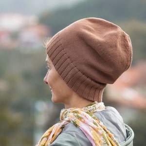 May include: A person wearing a brown knitted beanie hat with a scarf. The hat has a slouchy fit and is made of a soft, warm material.