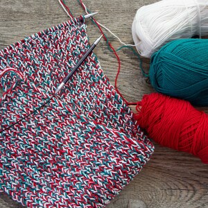 May include: A close-up of a knitted scarf in red, white, and green yarn. Three balls of yarn in the same colors are also visible.