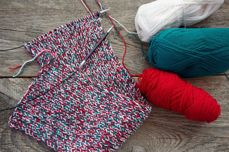 May include: A close-up of a knitted scarf in red, white, and green yarn. Three balls of yarn in the same colors are also visible.