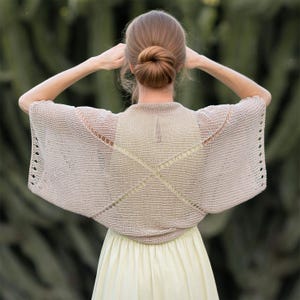 Könnte beinhalten: Ein beigefarbener Strickschal mit geometrischem Muster wird über einem gelben Kleid getragen. Der Schal hat eine quadratische Form am Rücken und erstreckt sich bis zu den Armen. Das Haar des Models ist zu einem Dutt gebunden.