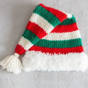 Op de afbeelding: Een rood, wit en groen gestreepte gebreide elfenmuts met een witte bontkraag. De muts heeft een witte pompon aan het uiteinde.