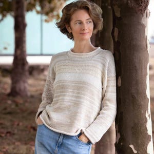 May include: A long-sleeved knit sweater featuring a horizontal striped pattern in shades of cream, beige, and light brown. The garment has a round neckline, a straight hem, and a textured knit construction, paired with light blue denim jeans.