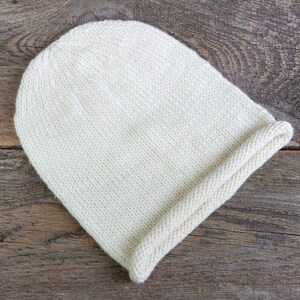 Peut inclure: Un bonnet en tricot blanc avec un bord replié. Le bonnet est fait d'un fil doux et texturé et est posé sur une surface en bois rustique.