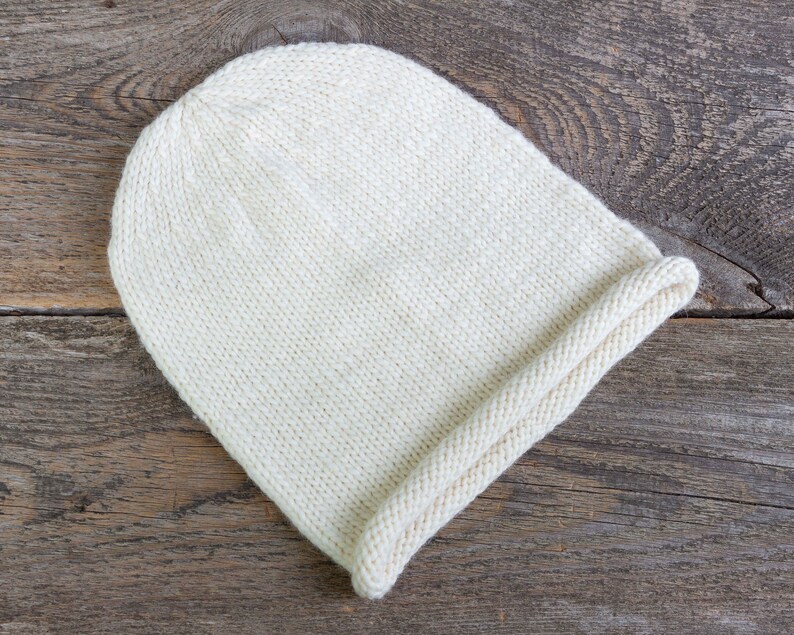 May include: A white knitted beanie hat with a folded brim. The hat is made of a soft, textured yarn and is lying on a rustic wooden surface.