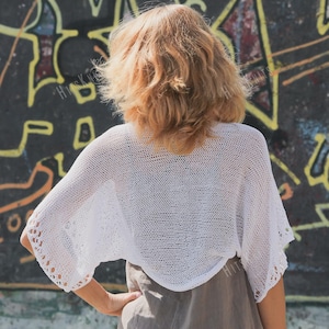 May include: A white, open-weave knit shrug with short sleeves. The shrug has a delicate, airy appearance, with a slightly cropped length. The background features a graffiti-covered wall with yellow and black paint.