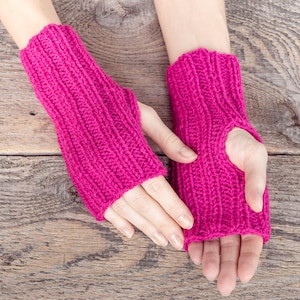 May include: A pair of pink knitted fingerless gloves with a ribbed texture. The gloves are shown on a wooden surface.