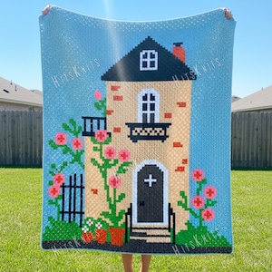 Op de afbeelding: Een lichtblauwe gebreide deken met een huisontwerp. Het huis heeft een zwart dak, een wit raam en een zwarte deur met een kruis. Roze bloemen en groen gebladerte versieren het huis. De tekst "Hits Knits" staat herhaald rond de randen.
