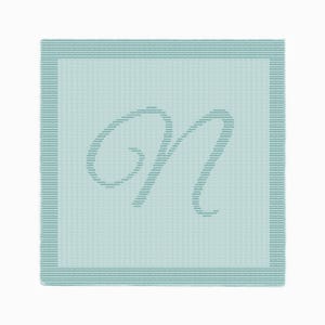 May include: A square, light blue blanket with a large, stylized letter "M" in the center. The letter and the border are a darker shade of blue. The blanket has a textured appearance.
