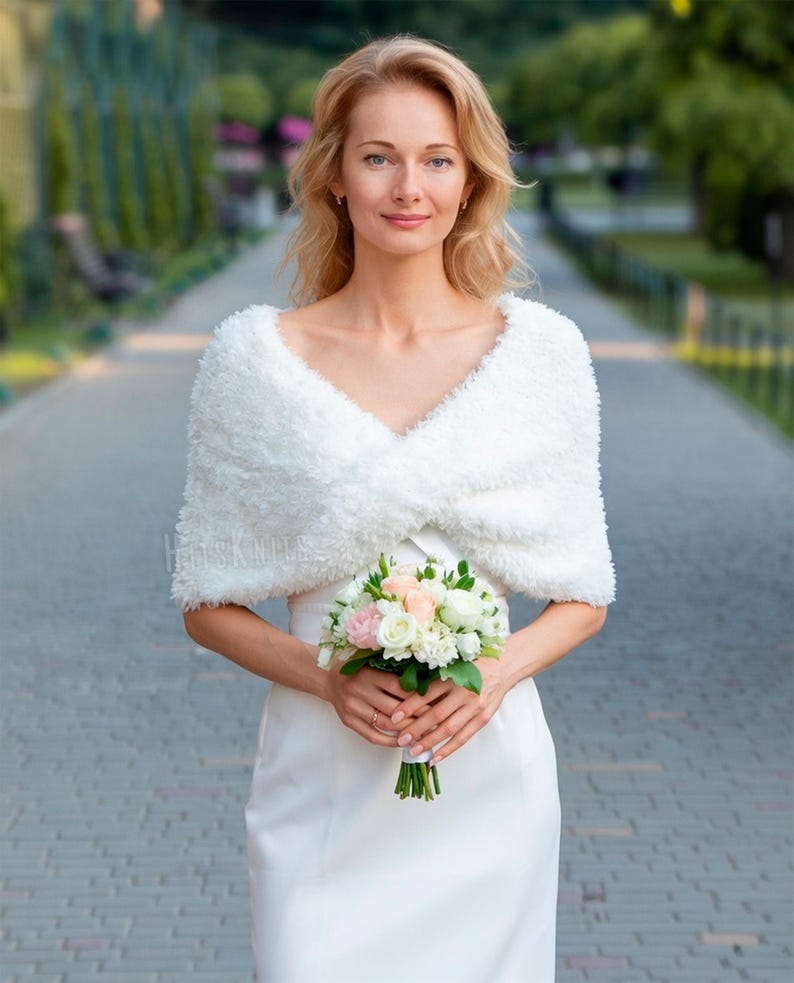 May include: A white faux fur shrug with a textured, fuzzy appearance and a V-neckline, worn over a plain white dress. The garment features short sleeves and a wrap-style front, providing a soft, layered look. A bouquet of white and pink roses is held.