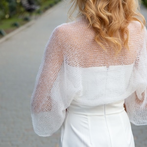 May include: White crochet shrug with a delicate open-weave pattern. The shrug has long sleeves and a loose fit.