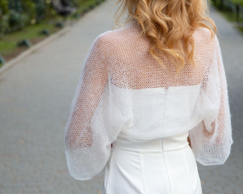May include: White crochet shrug with a delicate open-weave pattern. The shrug has long sleeves and a loose fit.