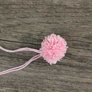 Puede incluir: Un pomp&oacute;n de lana rosa con una larga cola de lana rosa unida a &eacute;l. El pomp&oacute;n est&aacute; sobre una superficie de madera.