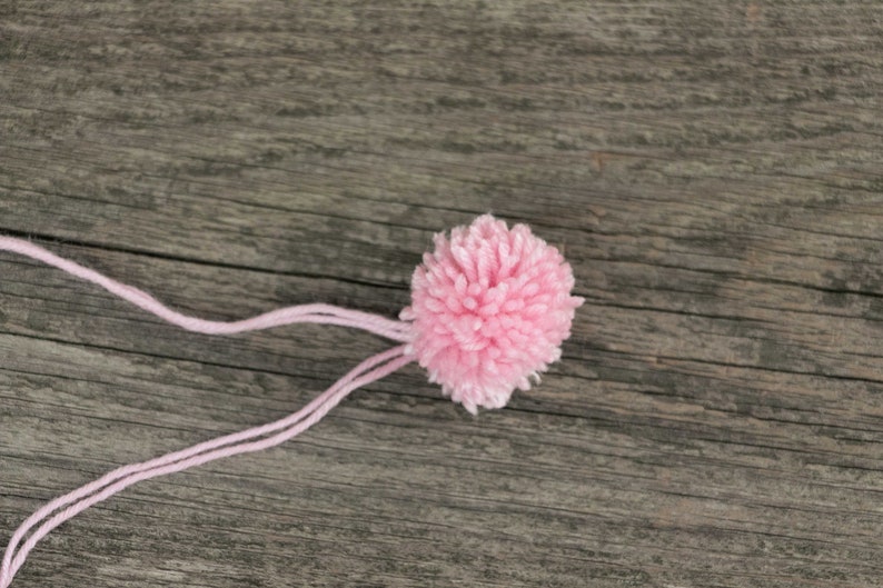 Puede incluir: Un pomp&oacute;n de lana rosa con una larga cola de lana rosa unida a &eacute;l. El pomp&oacute;n est&aacute; sobre una superficie de madera.
