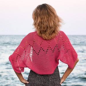 May include: A vibrant pink, open-weave knitted shawl with short sleeves. The shawl has a decorative pattern across the back and shoulders. The woman is standing in front of a body of water.