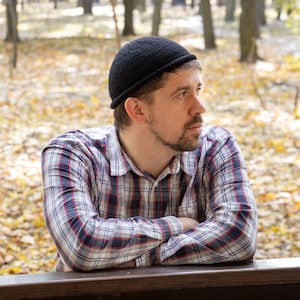 May include: A man wearing a black beanie and a plaid shirt with his arms crossed, standing in front of a wooden railing. The background is a blurred image of trees with yellow leaves.