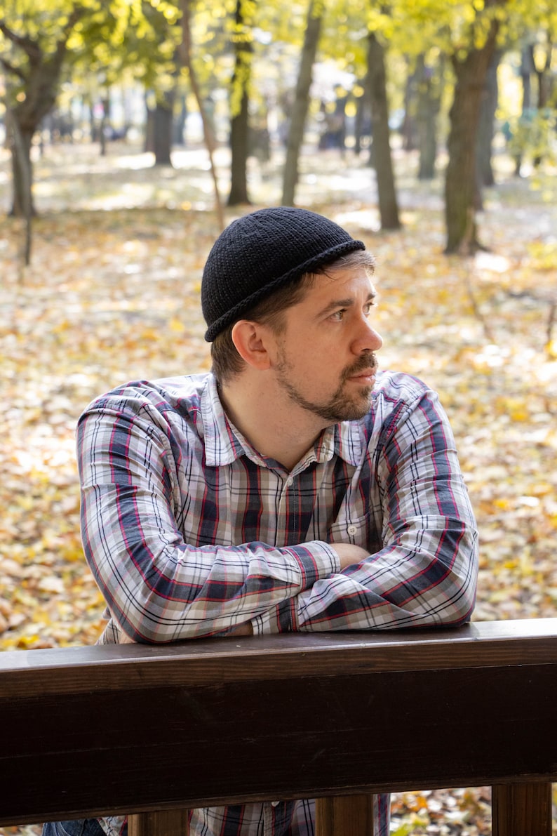 May include: A man wearing a black beanie and a plaid shirt with his arms crossed, standing in front of a wooden railing. The background is a blurred image of trees with yellow leaves.