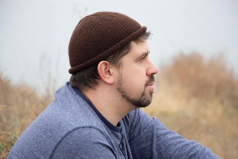 May include: A man wearing a brown knitted beanie and a blue long-sleeved shirt is looking off to the side.