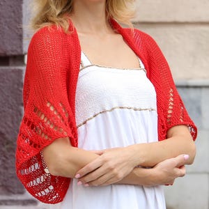K&ouml;nnte beinhalten: Ein leuchtend roter Strick-Bolero mit einem durchbrochenen Muster und zarten &Ouml;sendetails entlang der &Auml;rmel. Das Kleidungsst&uuml;ck hat ein lockeres, strukturiertes Gewebe und wird &uuml;ber einem wei&szlig;en &auml;rmellosen Oberteil mit gesmoktem Oberteil getragen.