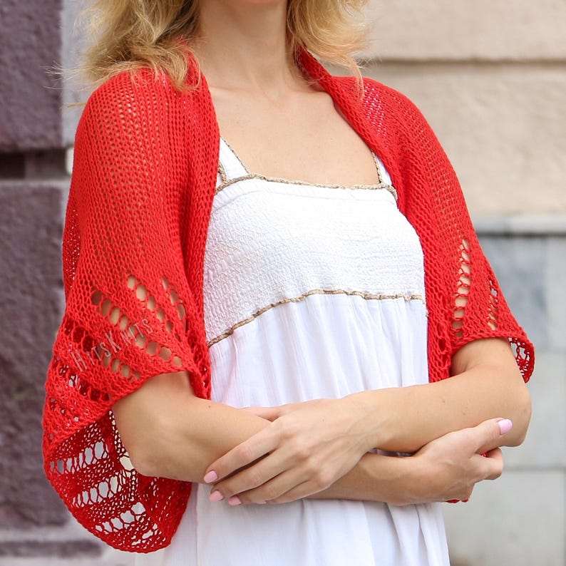 K&ouml;nnte beinhalten: Ein leuchtend roter Strick-Bolero mit einem durchbrochenen Muster und zarten &Ouml;sendetails entlang der &Auml;rmel. Das Kleidungsst&uuml;ck hat ein lockeres, strukturiertes Gewebe und wird &uuml;ber einem wei&szlig;en &auml;rmellosen Oberteil mit gesmoktem Oberteil getragen.