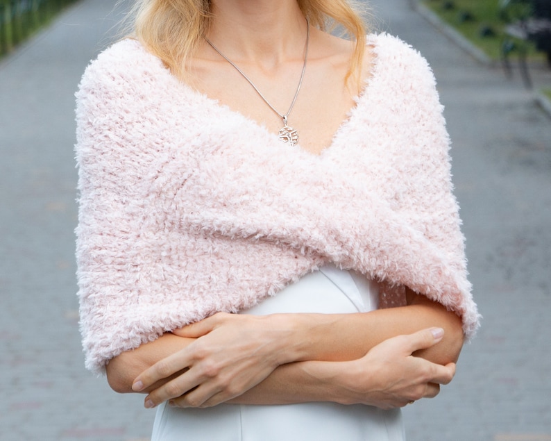 May include: A pale pink, fuzzy, textured knit shrug with a wrap-around design that covers the shoulders and upper arms. The garment features a soft, shaggy yarn construction with a V-neckline, worn over a plain white sleeveless garment.