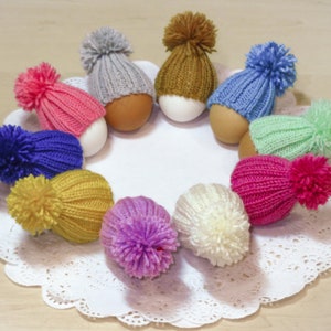 Könnte beinhalten: Eine Sammlung bunter Strickmützen mit Bommeln auf Eiern. Die Mützen sind in verschiedenen Farben, darunter rosa, blau, grün, gelb, lila und weiß.