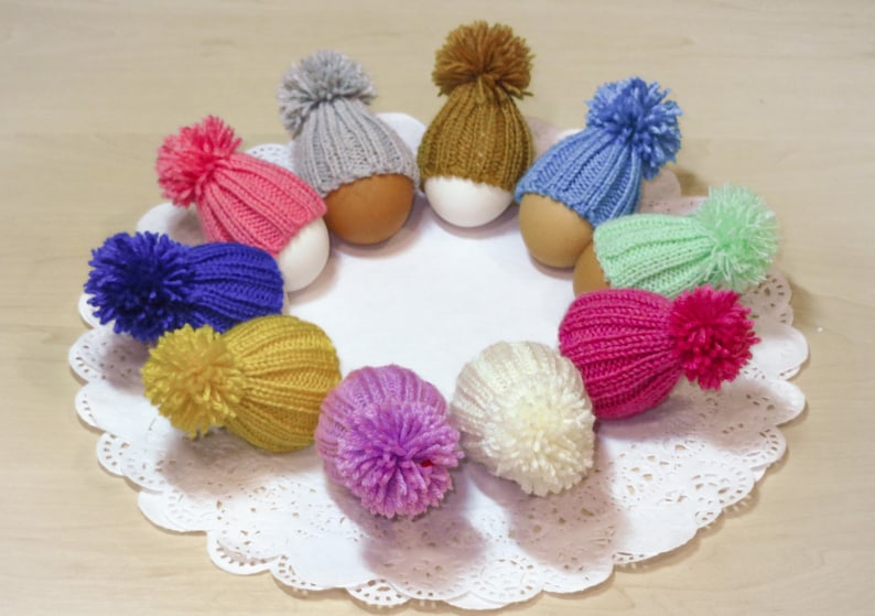 Puede incluir: Una colecci&oacute;n de gorros de punto coloridos con pompones en la parte superior de los huevos. Los gorros son de varios colores, incluyendo rosa, azul, verde, amarillo, morado y blanco.
