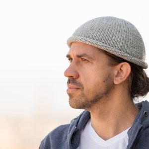 May include: A man wearing a gray knitted beanie with a long ponytail. He is wearing a gray hoodie and a white t-shirt.