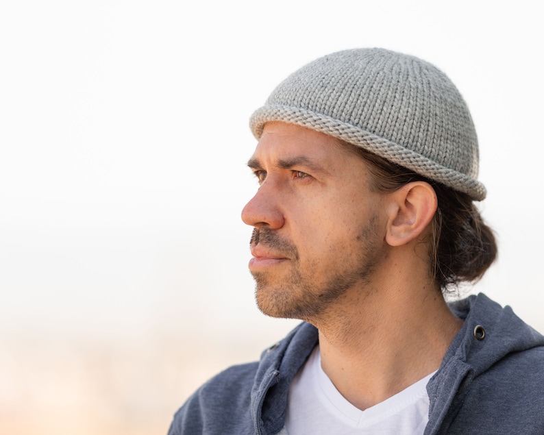 May include: A man wearing a gray knitted beanie with a long ponytail. He is wearing a gray hoodie and a white t-shirt.