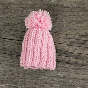 Puede incluir: Un gorro de punto rosa con un pomp&oacute;n esponjoso en la parte superior. El gorro est&aacute; sobre una superficie de madera.