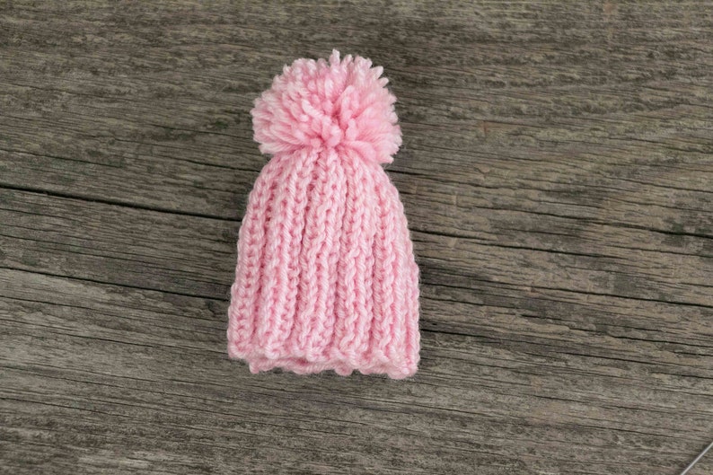 Puede incluir: Un gorro de punto rosa con un pomp&oacute;n esponjoso en la parte superior. El gorro est&aacute; sobre una superficie de madera.
