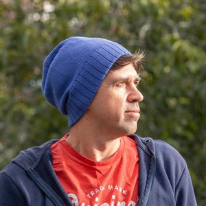 May include: A man wearing a blue beanie and a navy blue jacket over a red t-shirt with the text "TRADE MARK" printed on it.