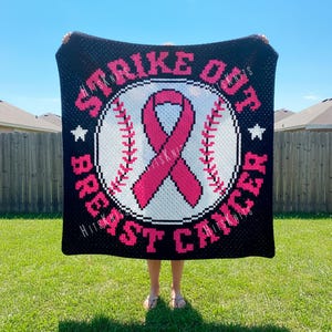 May include: Black blanket with a baseball design and the words "Strike Out Breast Cancer." The baseball has a pink ribbon in the center. White stars are on either side of the baseball. The blanket is held up in front of a green lawn.