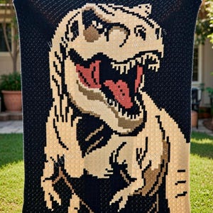 May include: A large, handmade crochet blanket featuring a pixelated Tyrannosaurus Rex design. The dinosaur is depicted in shades of tan, brown, and red against a black background. The blanket is held up, showcasing its size and intricate detail.