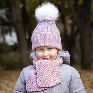 May include: A young girl wearing a pink and white knit hat with a white pom pom and a pink and white knit scarf. She is wearing a gray jacket and is smiling.