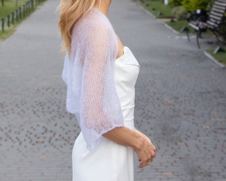 May include: A delicate, light purple knitted shawl with a lacy pattern. The shawl is draped over the shoulders of a person wearing a white dress.