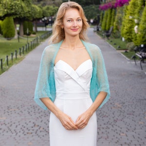 May include: A woman wearing a white dress with a light blue knitted shawl. She is standing on a paved walkway with green trees and bushes in the background.