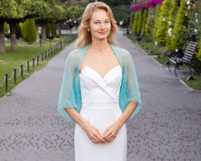 May include: A woman wearing a white dress with a light blue knitted shawl. She is standing on a paved walkway with green trees and bushes in the background.