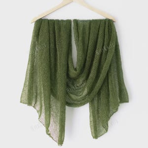 May include: Olive green knitted scarf displayed on a wooden hanger. The scarf is made of a lightweight, open-weave material, creating a delicate, airy appearance. The scarf is draped in a flowing style, showcasing its texture and color. The scarf is a versatile accessory.