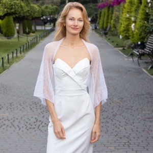 May include: A woman wearing a white dress with a delicate white lace shawl. She is standing on a paved path in a garden setting.