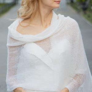 May include: A white, open-weave knitted shawl. The shawl is draped over the shoulders, creating a soft, flowing effect. The delicate fabric is light and airy. The garment is designed to be worn over the shoulders.