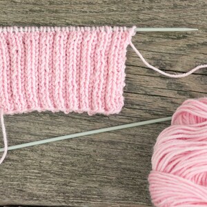 Puede incluir: Un ovillo de hilo rosa con una muestra de tejido de punto rosa en agujas de tejer. La muestra est&aacute; sobre una superficie de madera.
